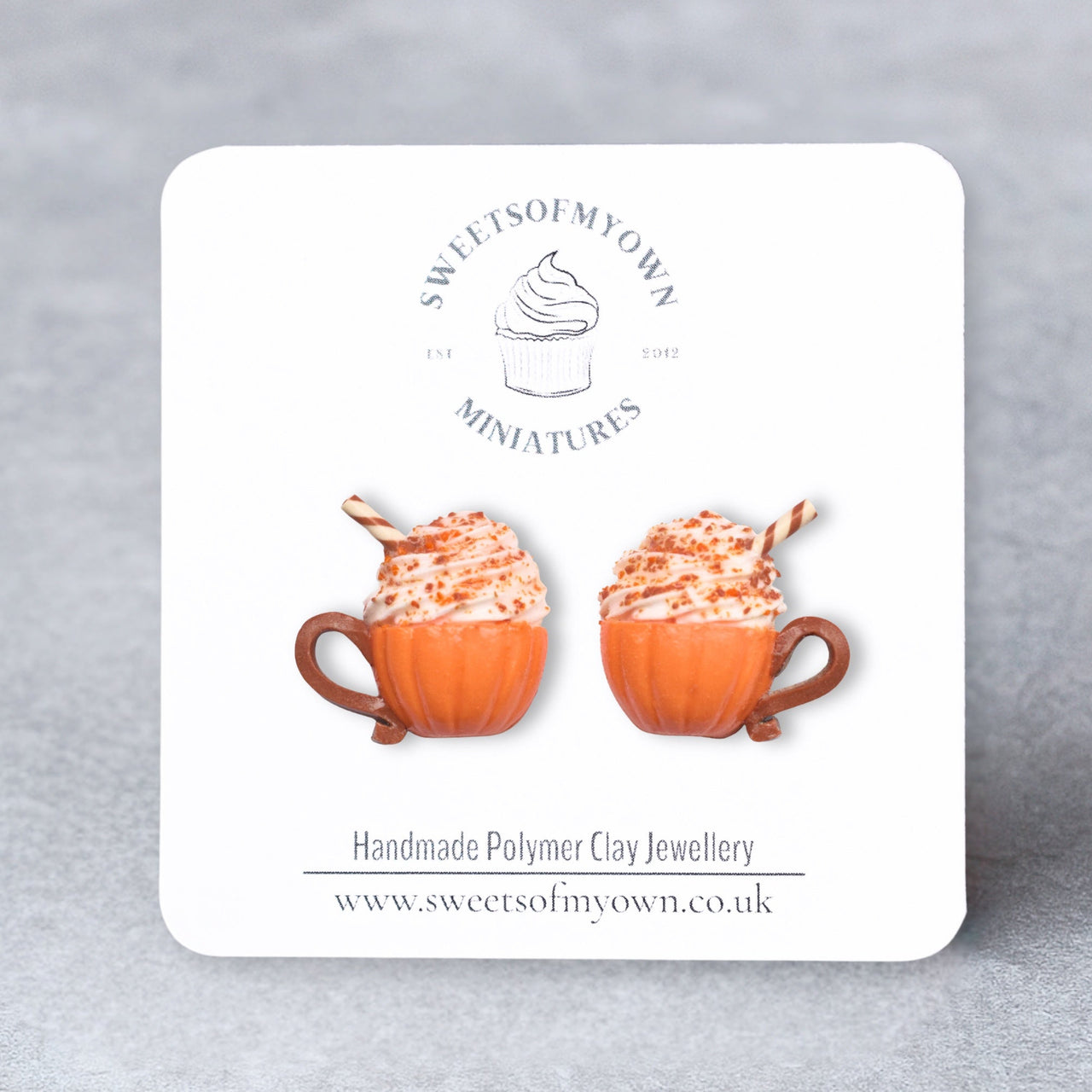 Card with polymer clay earrings resembling pumpkin spice lattes on a gray background