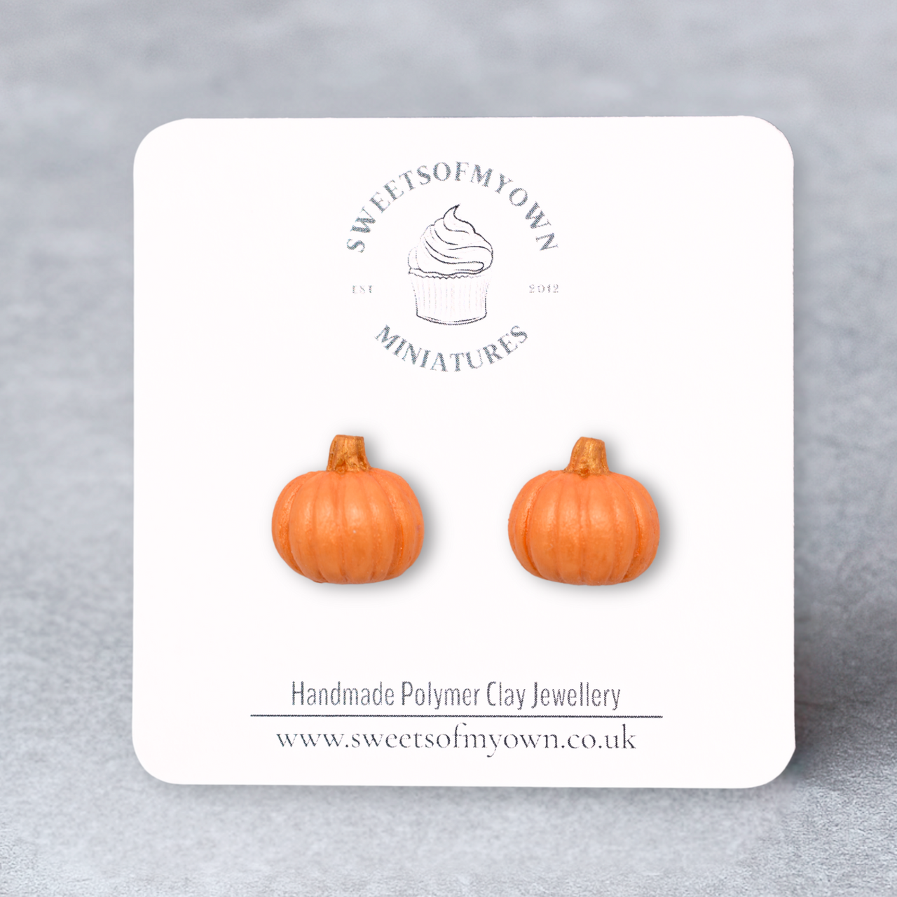 Pumpkin-shaped orange earrings on a branded card with a gray background
