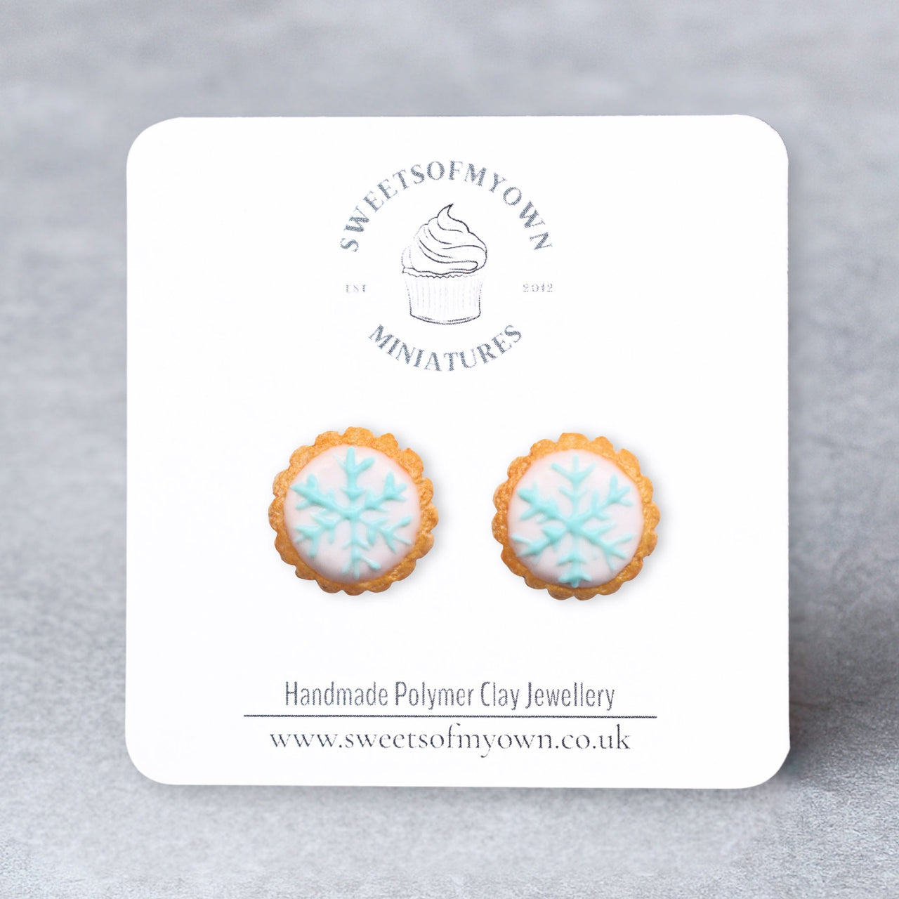 Pair of cookie-shaped earrings with white icing and blue snowflake on a Sweetsofmyown card against a gray background